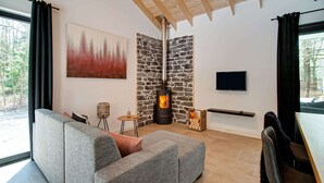 A modern living room with a stone fireplace, a grey sofa, a television, and a view of the outdoors. - Holiday Home in Biron With Barrel Sauna (Erezee)