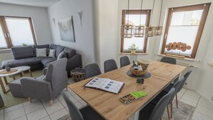 A modern living room with a wooden dining table, grey sofas, and a large window with blinds.