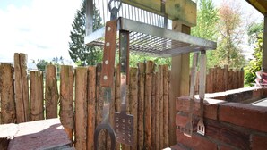 Un espace de cuisson extérieur rustique, doté d’un four en briques, d’une clôture en bois et de divers ustensiles de cuisine suspendus à un présentoir en métal.