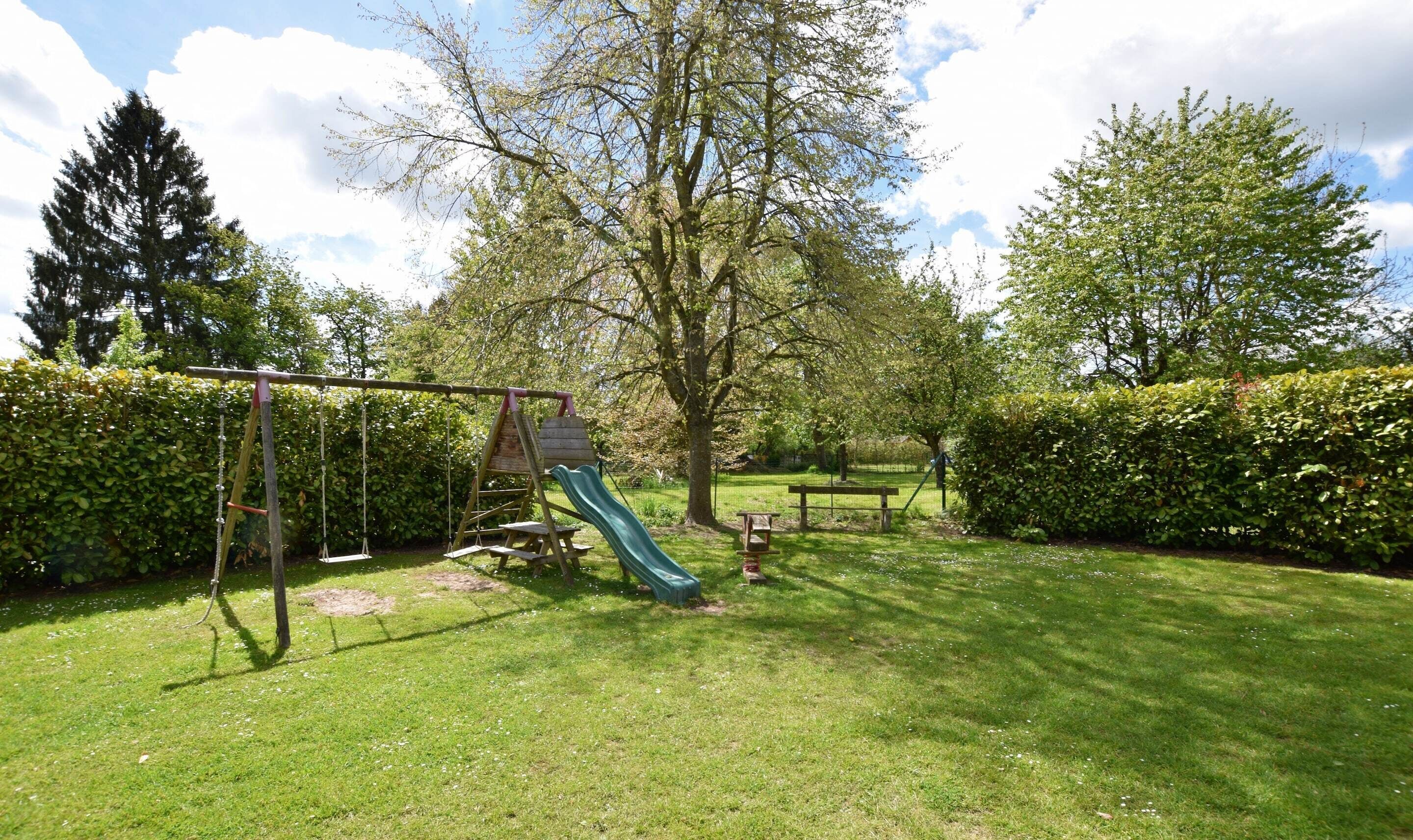 A playground with swings, a slide, and a climbing structure surrounded by well-maintained grass and hedges.