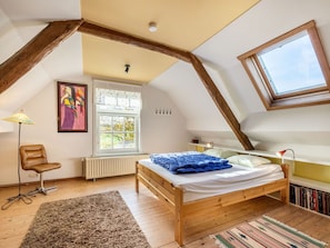 Ein Schlafzimmer mit schrägem Dach, einem hölzernen Bett, einem Heizkörper, einem Fenster, einem Stuhl und einem Bücherregal.