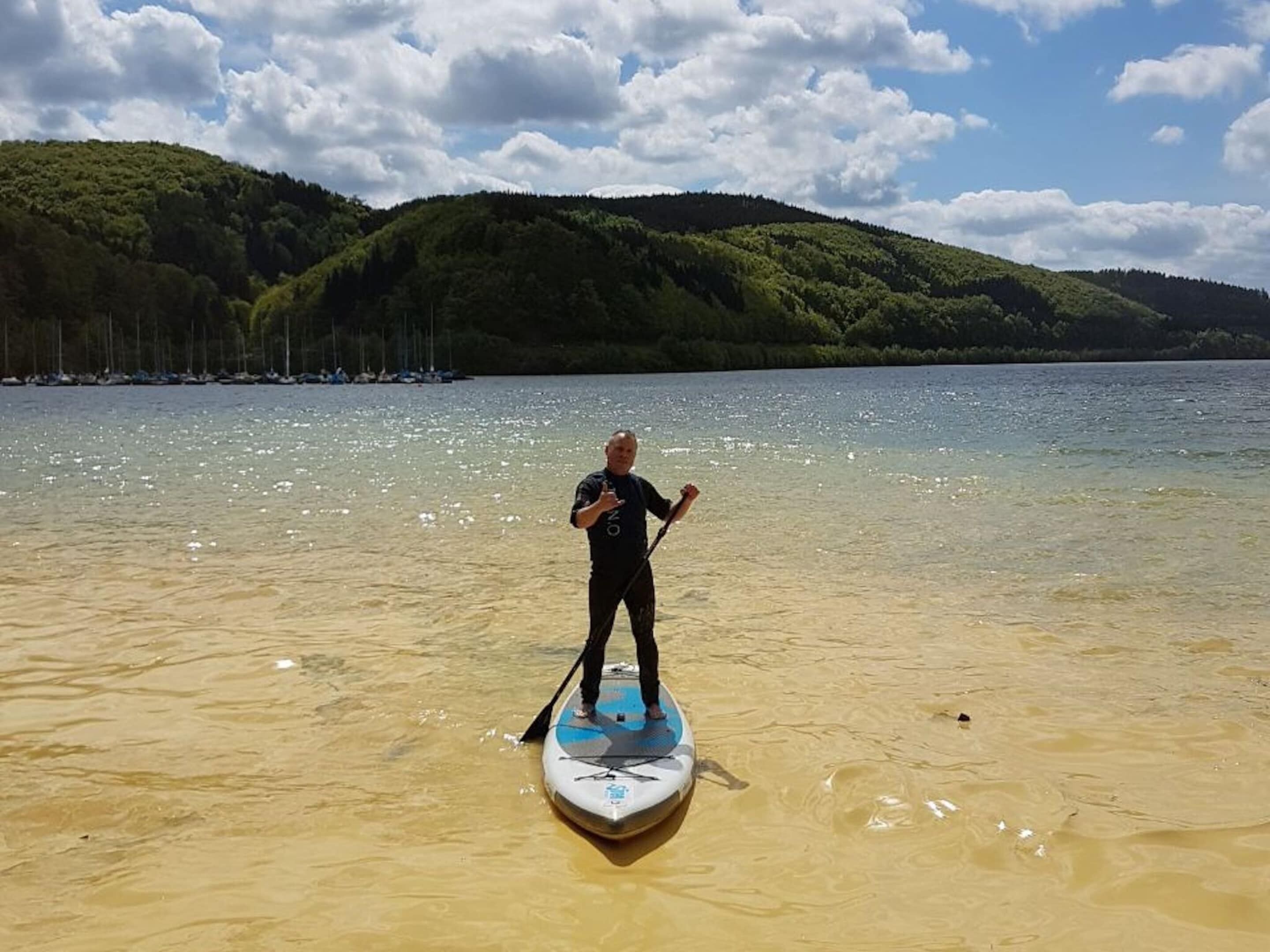 Eine Person steht auf einem Paddleboard im Wasser, im Hintergrund ein bewaldeter Hügel.