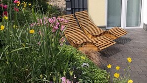 Una silla de madera para relajarse en un jardín con diversas flores y un edificio al fondo.