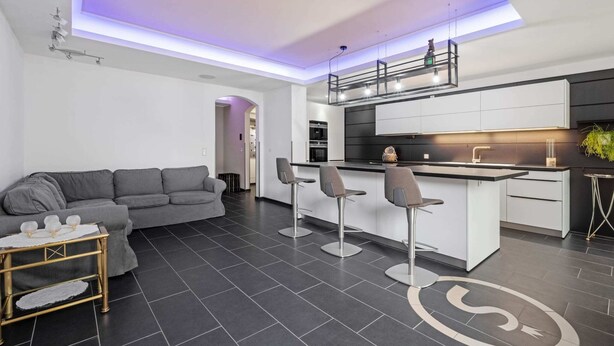 Una cocina moderna con una isla central, taburetes de bar y un sofá gris en la sala de estar.