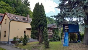 A residential area with a yellow house, a tall coniferous tree, a blue slide, and a wooden playhouse. - Great Holiday Home With Terrace Near Saxon (Hohnstein)