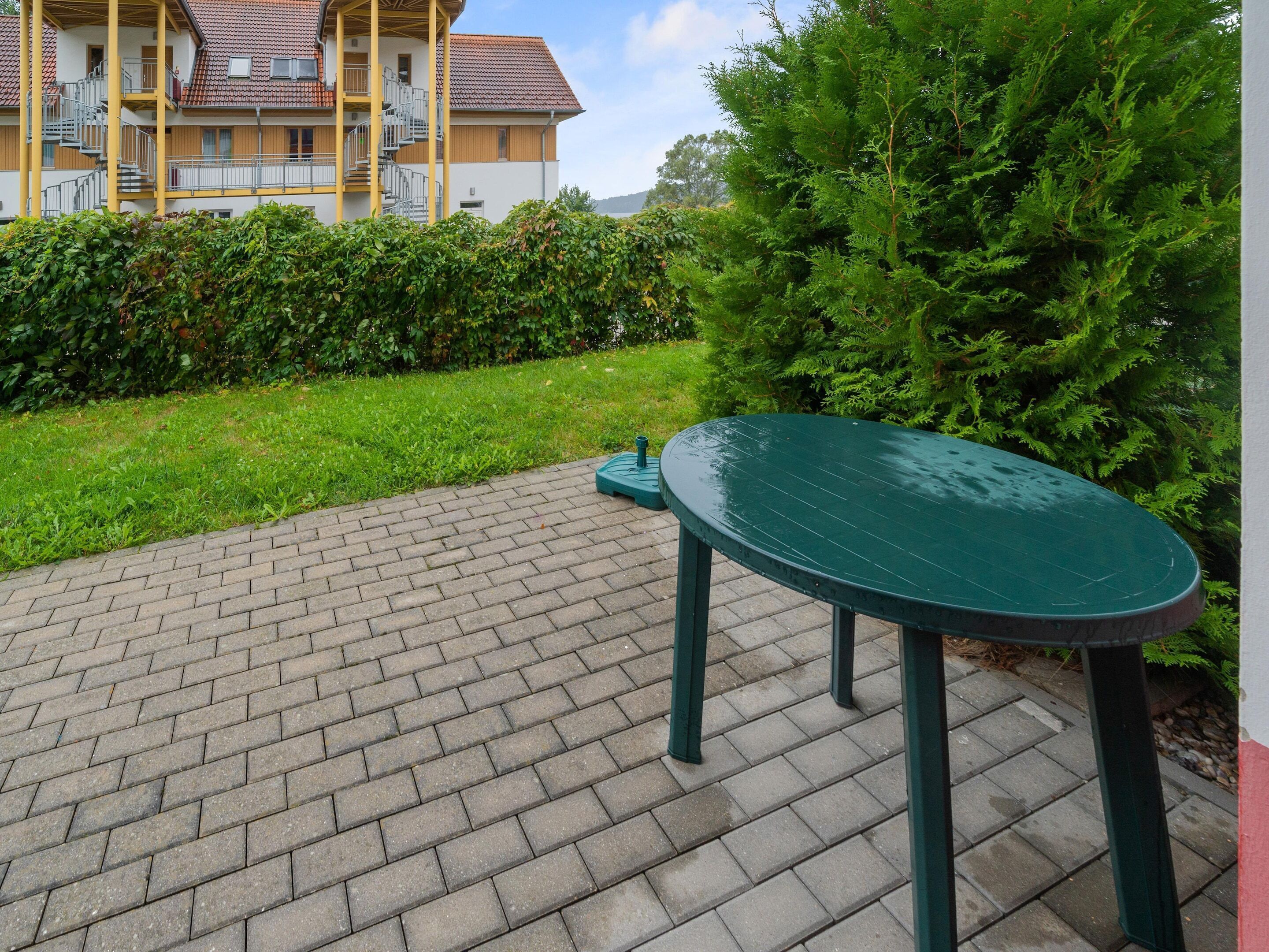 A paved outdoor area with a green table and chairs, surrounded by greenery and a building with balconies.