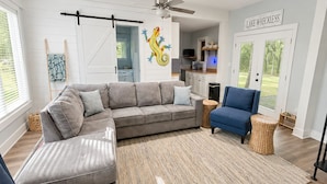 A modern living room with a sectional sofa, a blue armchair, a woven rug, and a ceiling fan. - Lake Murray Cottage w/ Private Dock, Fire Pit & Kayaks - Near Martin's Landing (Prosperity)