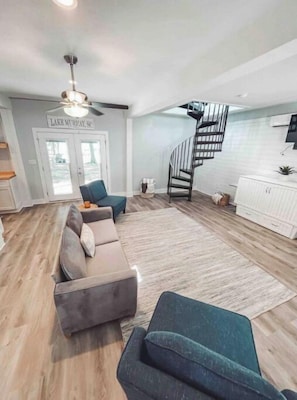 A spacious living room with a grey sectional sofa, a teal armchair, and a wooden floor. A staircase leads to a kitchen area with white cabinets and a white fridge.