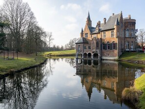 Un bâtiment historique entouré de douves, dont l’image se reflète dans l’eau, ainsi que celle de la structure et des arbres environnants.