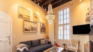 Un salón acogedor con un sofá, una mesa de centro y elementos decorativos. Se aprecia un ventanal con vistas, una araña y obras de arte en las paredes.