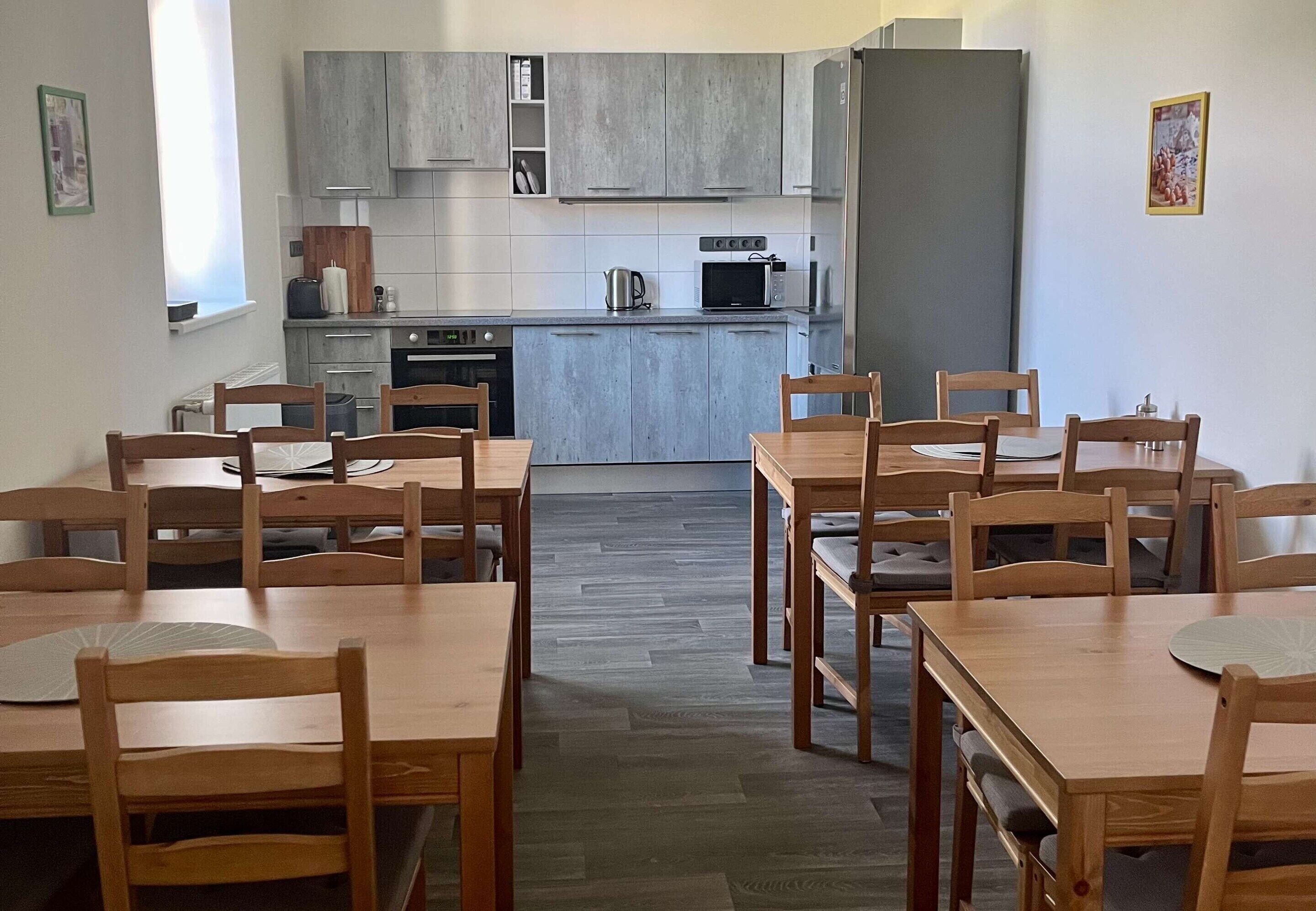 Una cocina moderna con armarios grises, un microondas y una tetera. Se observan mesas y sillas de madera dispuestas al frente.