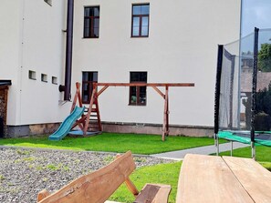 Et legetårn i træ med gyngestol og rutchebane i en baghave, med en hvid bygning i baggrunden.