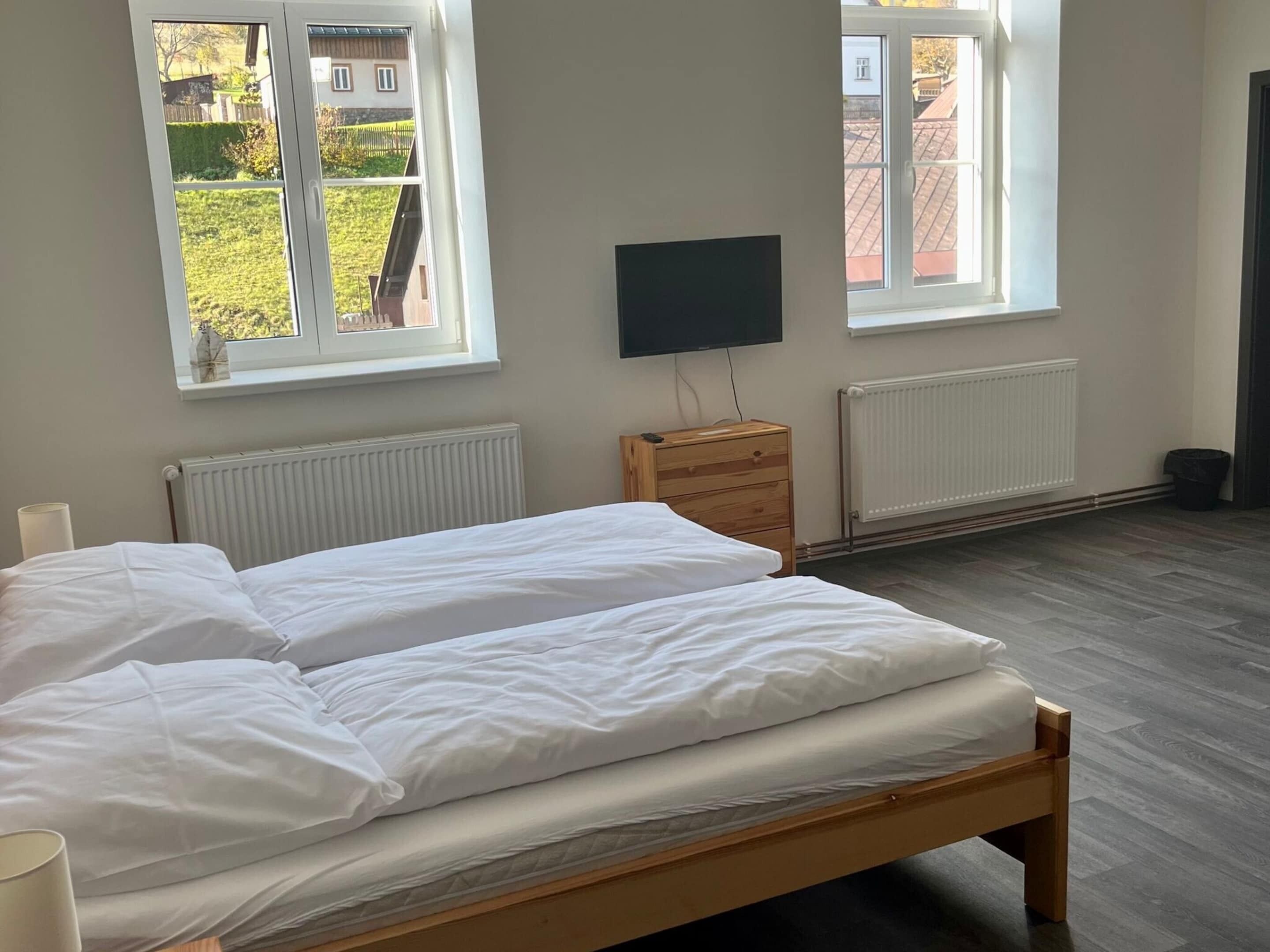 A bedroom with two beds, a wooden dresser, a TV, and two windows with a view of the outside.