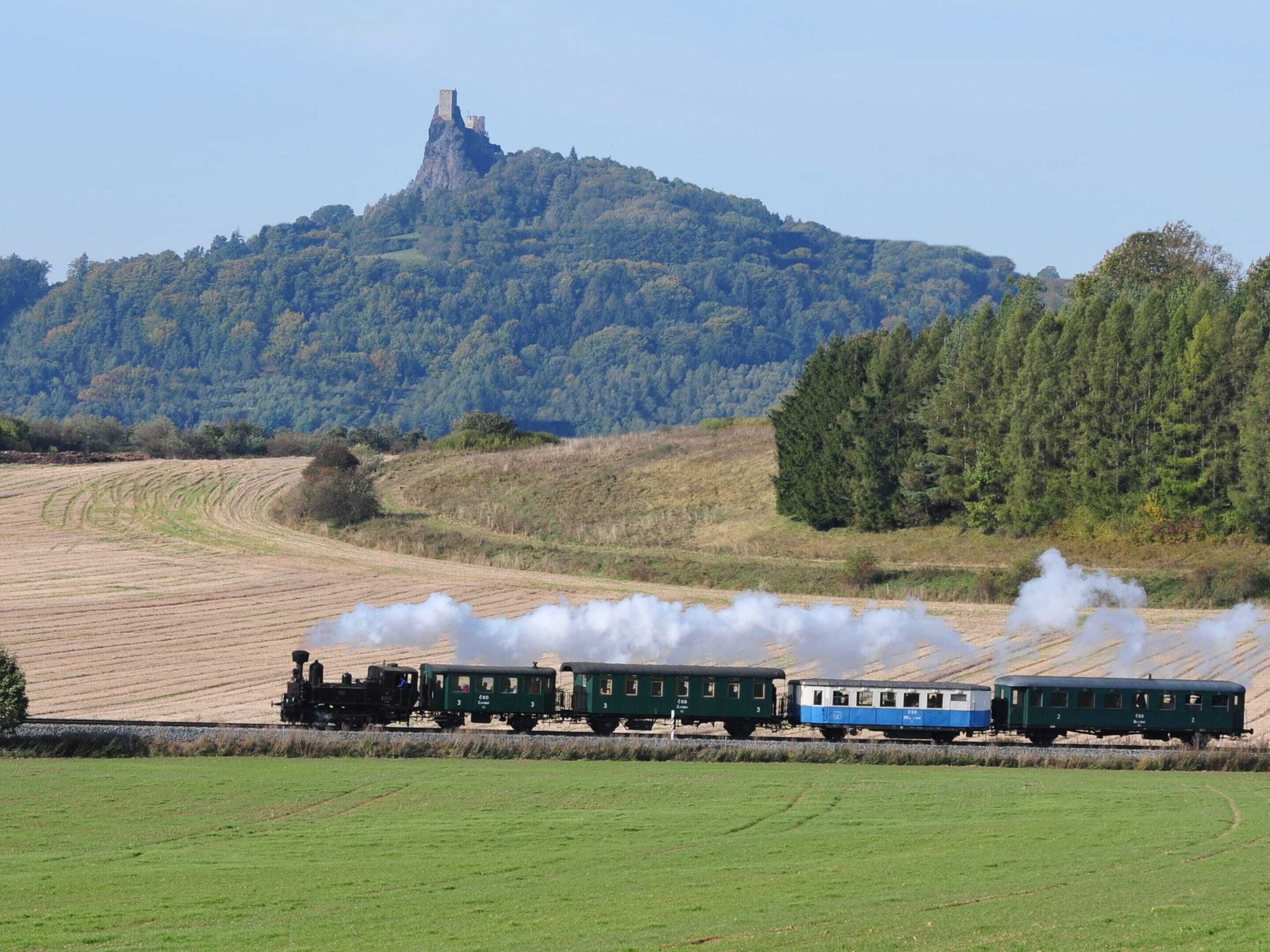 Un train à vapeur, avec des wagons verts et bleus, traverse un paysage rural composé de champs et d’une colline sur laquelle se dresse une structure ressemblant à un château, visible au loin.