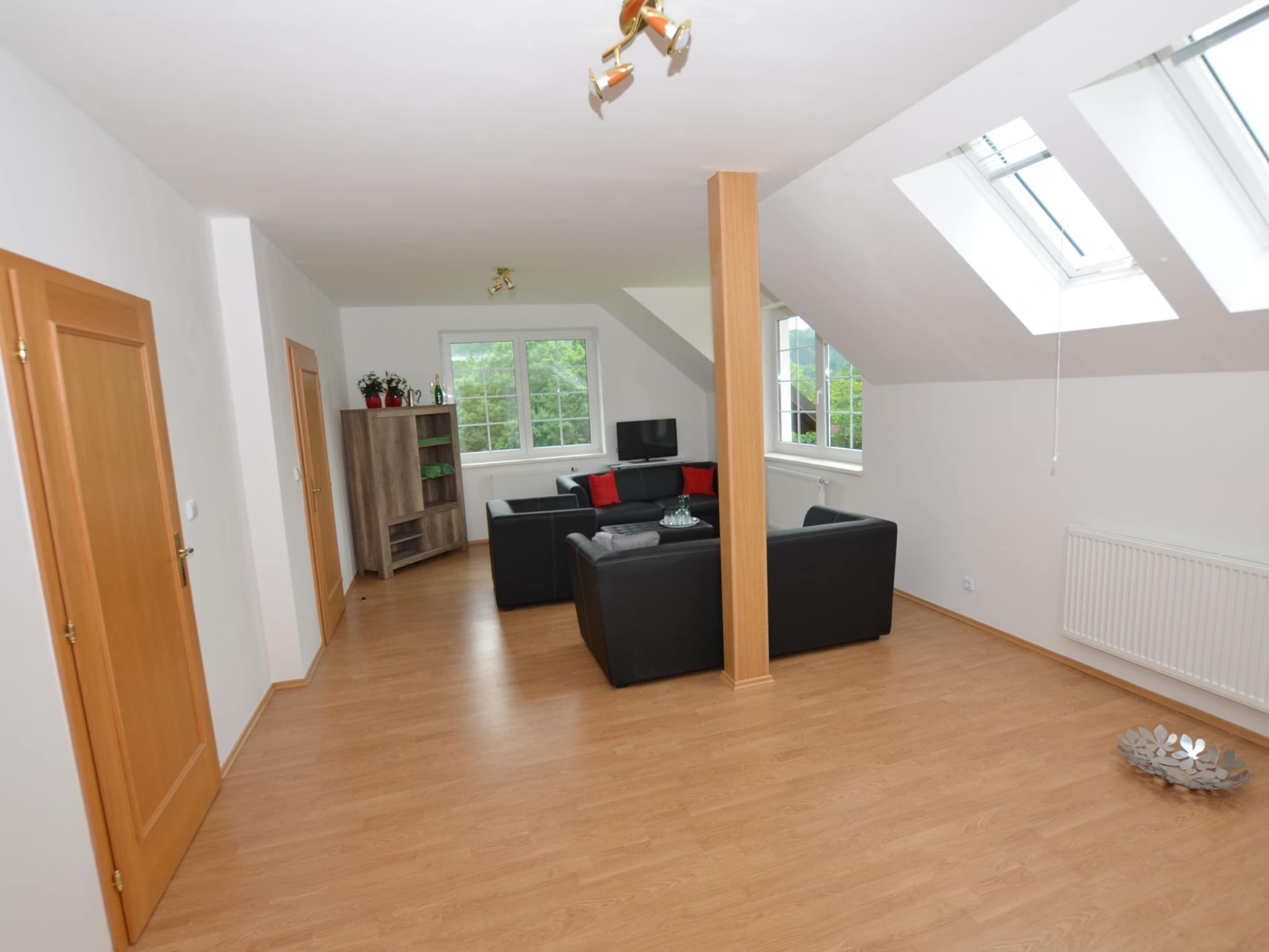 A spacious living area with a high ceiling, wooden flooring, and a built-in bookshelf. There are two black sofas and a small table with a decorative bowl.