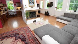 A spacious living room with a grey sofa, a fireplace, a TV, and a large window with a view of greenery. - Tasteful Villa in Zernov With Private Pool (Žernov)