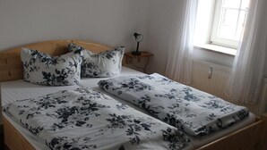 Uma cama de casal com roupa de cama floral, uma cabeceira de madeira, uma mesa de cabeceira com abajur e uma janela com cortinas.