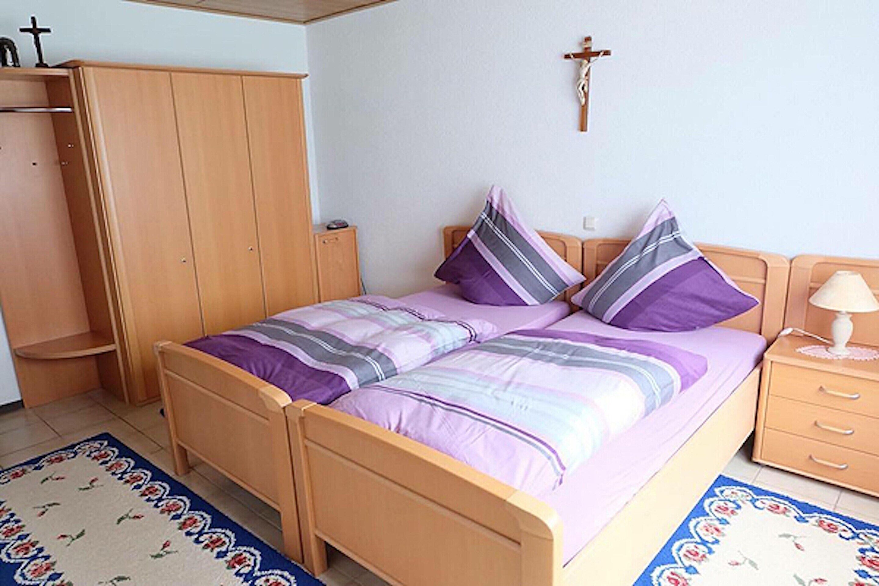 A bedroom with a wooden bed, two pillows, a nightstand, a lamp, a wardrobe, and a cross on the wall.