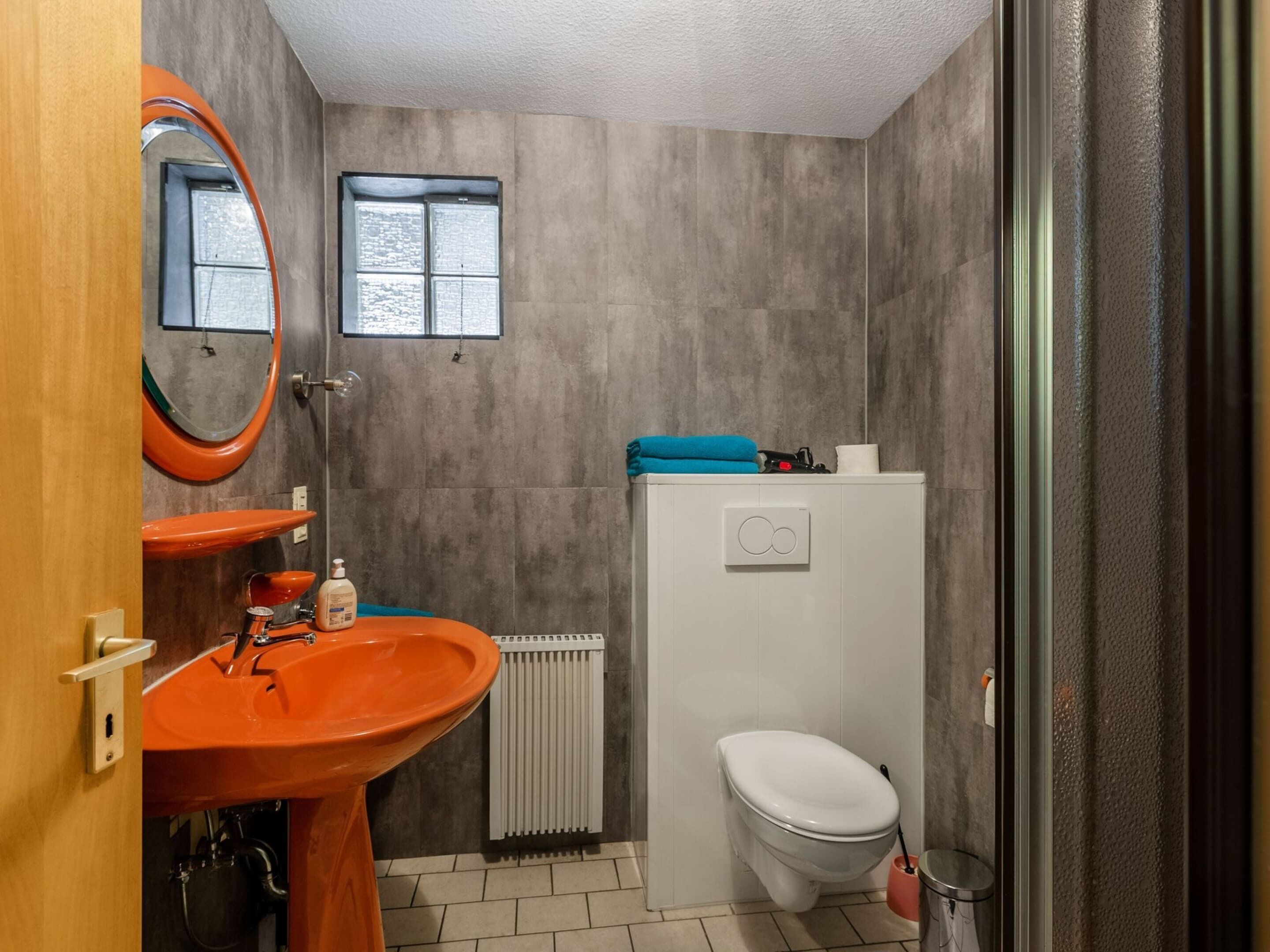 A bathroom with a wooden door, an orange sink, a white toilet, and a window above the toilet.