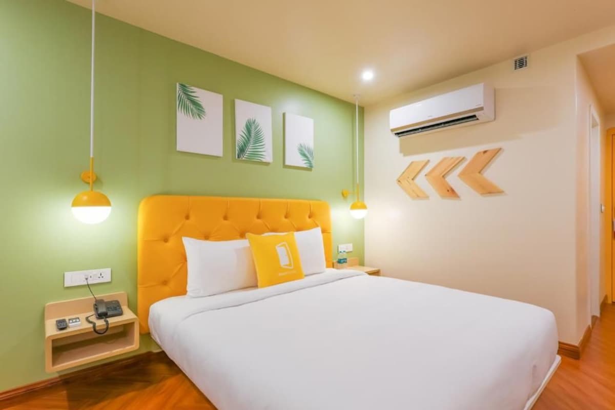 A hotel room with a bed, a nightstand, an air conditioning unit, and wall-mounted artwork.