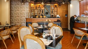 A restaurant interior with wooden flooring, high ceilings with black railings, and a central bar area with a person working at the counter. - Bloom Hotel - Golf Course Road, Sector 43 (Gurugram)