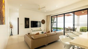 A modern living room with a beige sofa, a flat-screen TV, and a dining area with white chairs and a table. - Oriental 3br Pool Villa (Rawai)