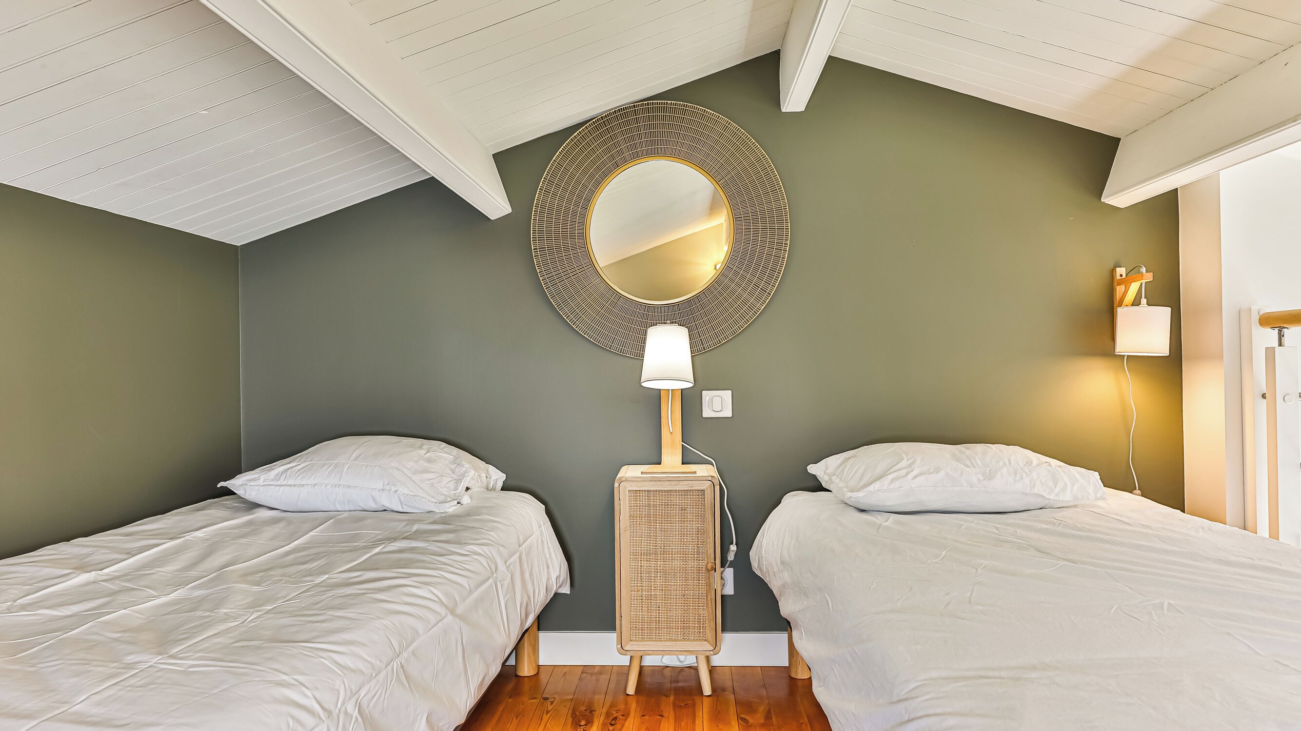 A room with two single beds, a round mirror, a lamp, and a rattan speaker.