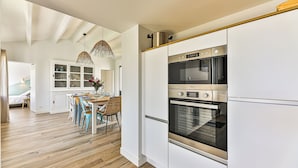 A modern kitchen with white cabinetry, a stainless steel oven, and a dining area with a wooden table and chairs. - Noirmoutier-le vieil Charming house with enclosed garden 300m from the beach (Noirmoutier-en-l'Île)