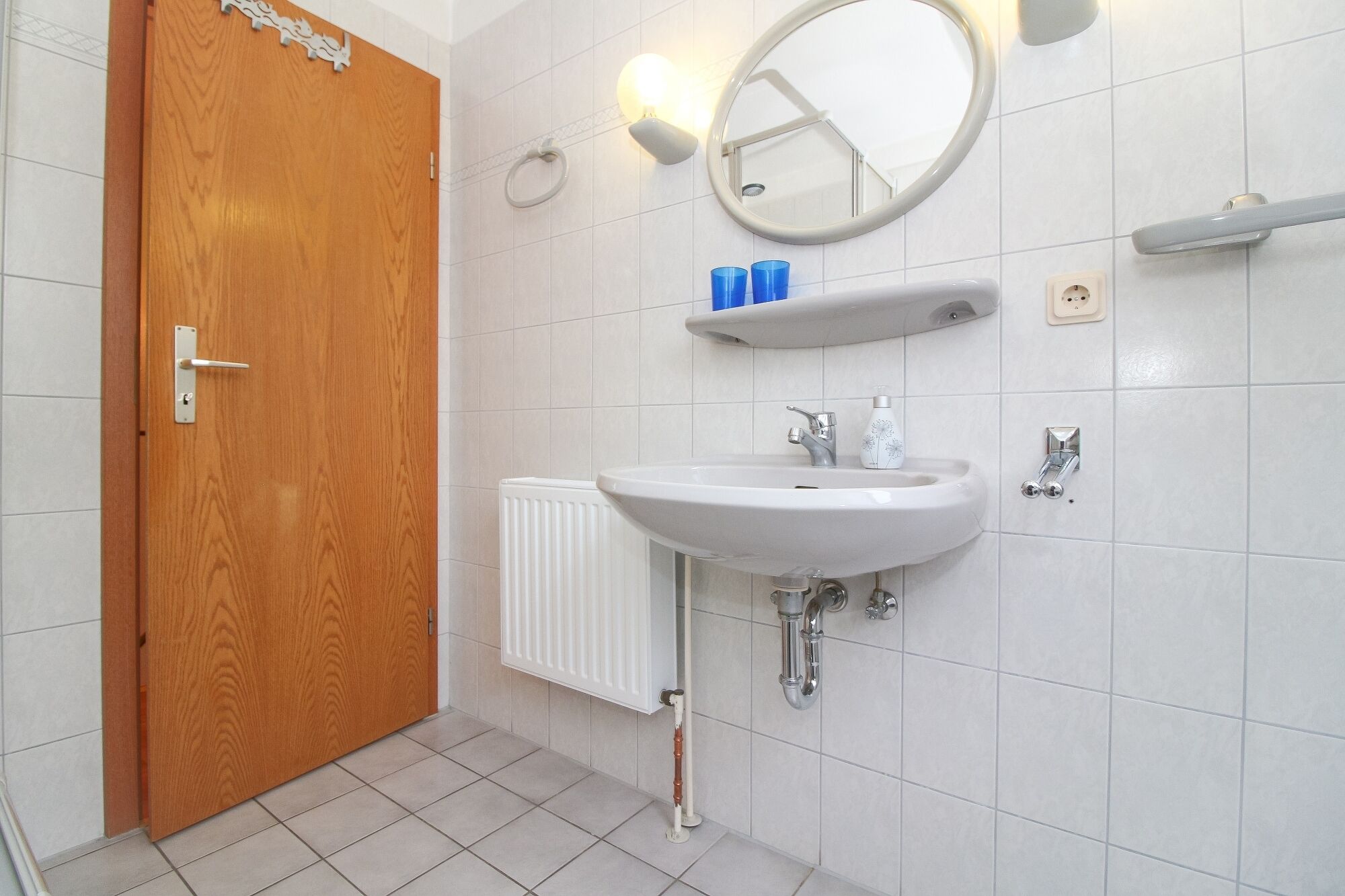 Un bagno con porta in legno, pareti piastrellate di bianco e un lavandino bianco con uno specchio sopra, oltre a un porta asciugamani.