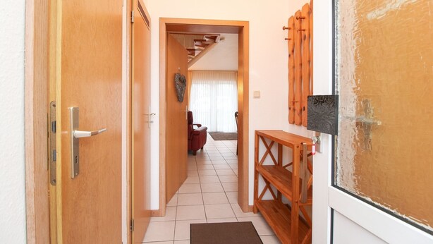 Een gang met houten deuren, een bankje en een doorkijk naar een woonkamer met een bank en gordijnen.