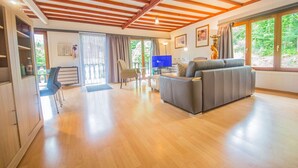 A spacious living area with a wooden ceiling, a grey sofa, a television, and a large window with a view of greenery. - Chalet in Rendeux With Garden (Rendeux)