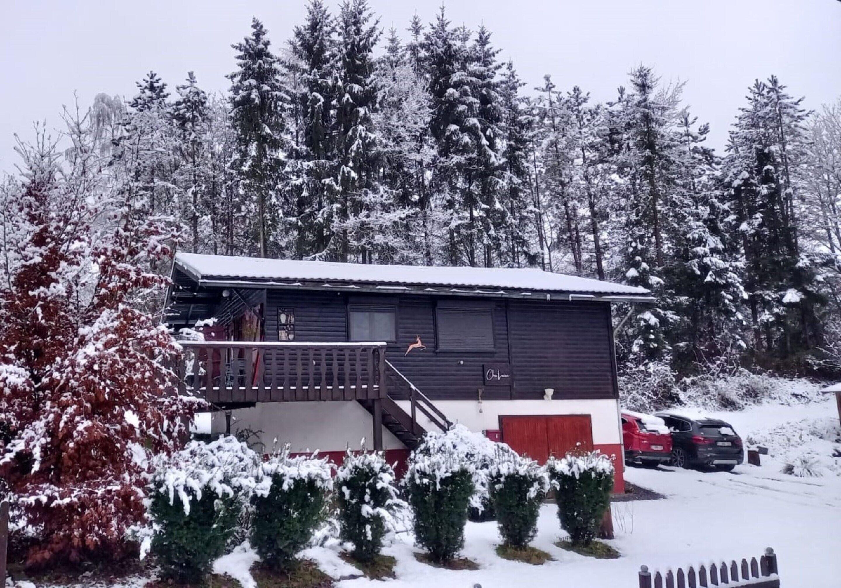 A cabin with a balcony, surrounded by snow-covered trees and a red car parked in the driveway.
