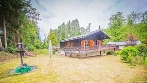 A cabin with a wooden deck, surrounded by a grassy yard and dense forest. - Chalet in Rendeux With Garden (Rendeux)