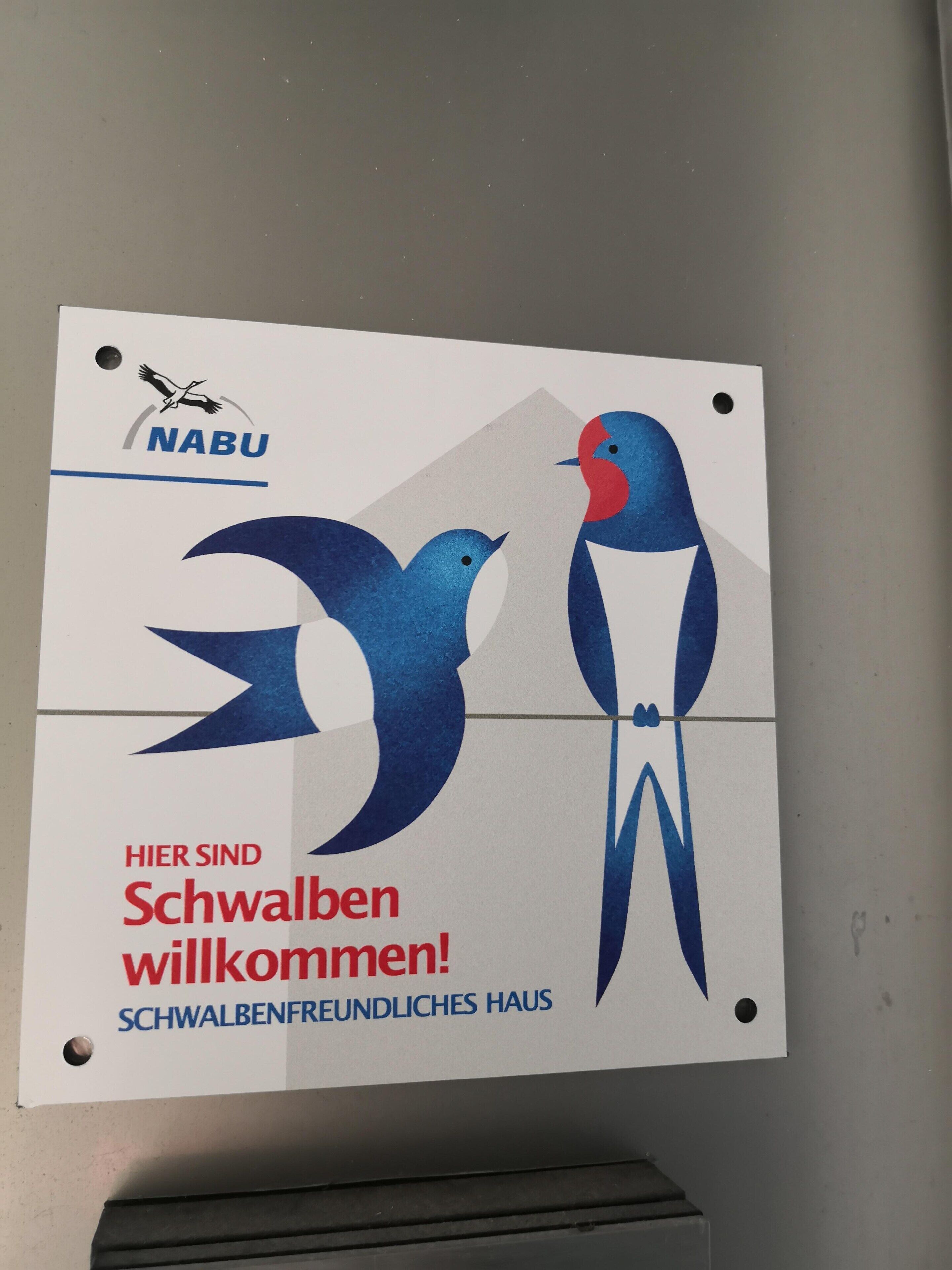 Ein Schild mit zwei stilisierter Vögel und Text, der eine freundliche Botschaft für Schwaben vermittelt.