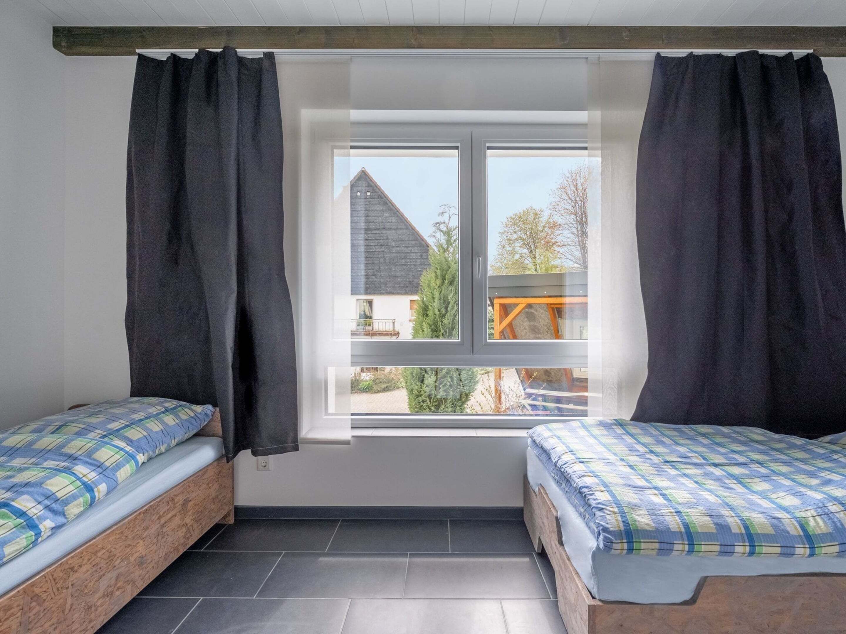 A room with two beds, a window with curtains, and a view of a house and trees outside.