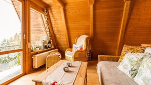 A cozy attic bedroom with wooden walls and a sloped ceiling. It features a bed, a wicker chair, a small table with a lamp, and a window offering a view of the outdoors. - Gemutliches Waldversteck in Suhl (Suhl)
