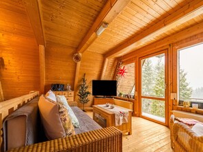 Interior acogedor de una cabaña con techo de madera, un sofá con cojines, un pequeño árbol de Navidad, un televisor y un amplio ventanal con vistas a árboles.
