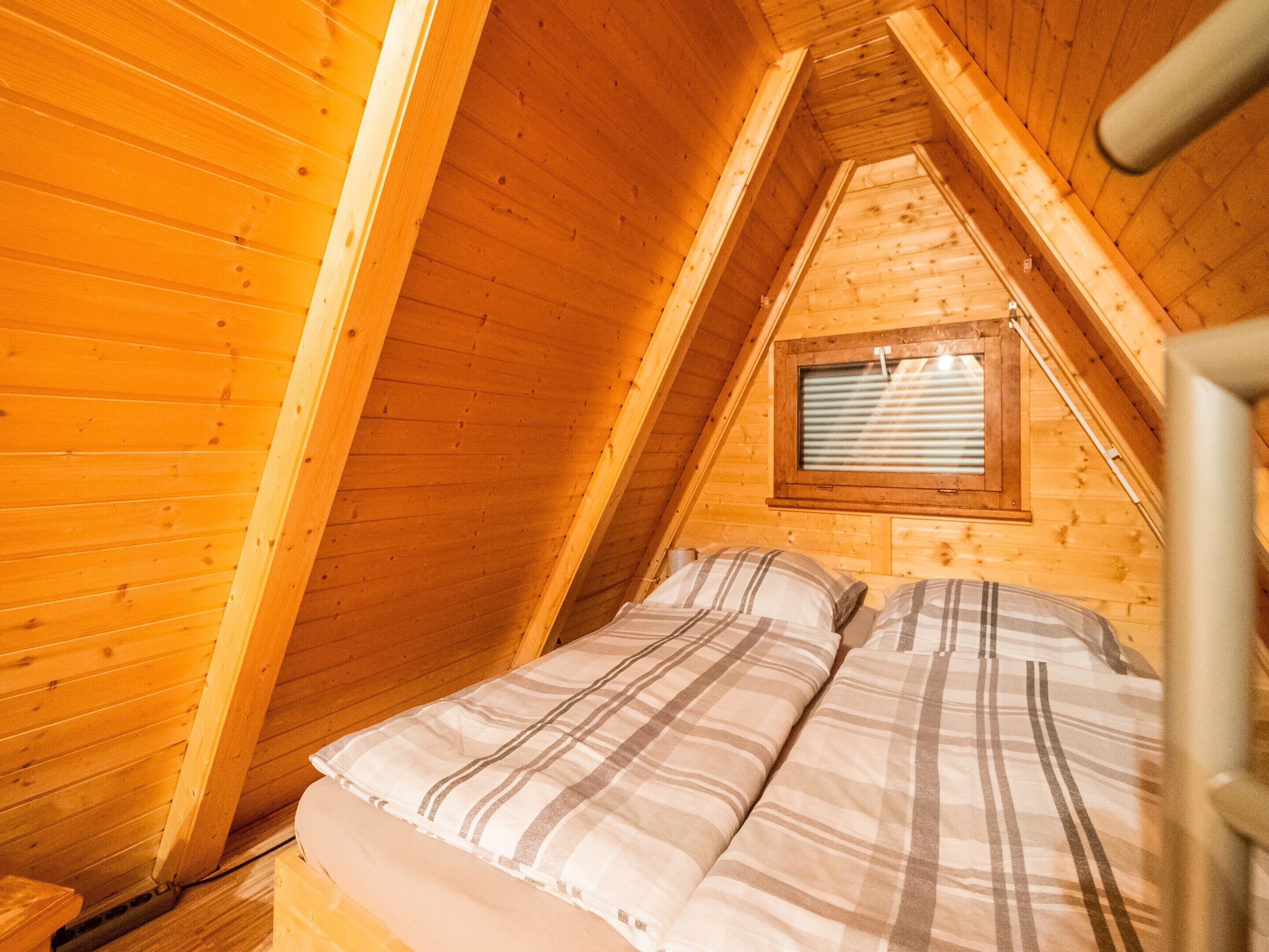 A small, cozy attic bedroom with wooden walls and a sloped ceiling. There is a bed with plaid bedding and a window with blinds.