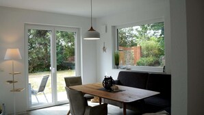 Ein Essbereich mit Holztisch, vier Stühlen, einer Couch, einer Pendelleuchte und Blick auf einen Garten durch große Fenster.