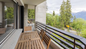A balcony with a wooden table and chairs, a concrete pillar, and a view of trees and mountains.