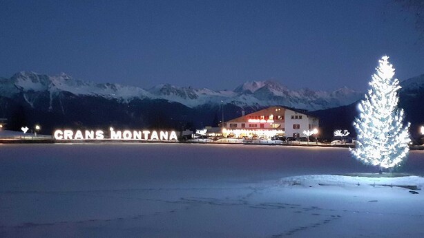 A building with illuminated signage reading "GRANS MONTANA" and a snow-covered landscape with mountains in the background.