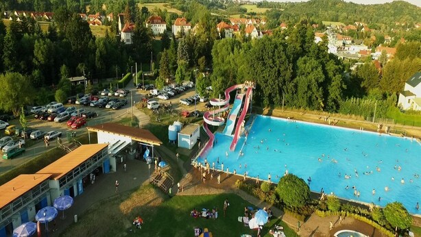 An aerial view of a swimming pool complex with a water slide, surrounded by parked cars and a building with outdoor seating.