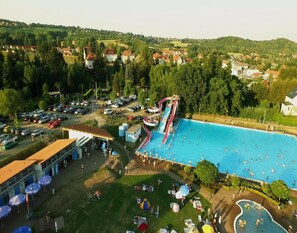 Vue aérienne d’un complexe de piscines doté d’un toboggan aquatique, entouré de voitures garées et d’un bâtiment avec des espaces de repos en plein air.