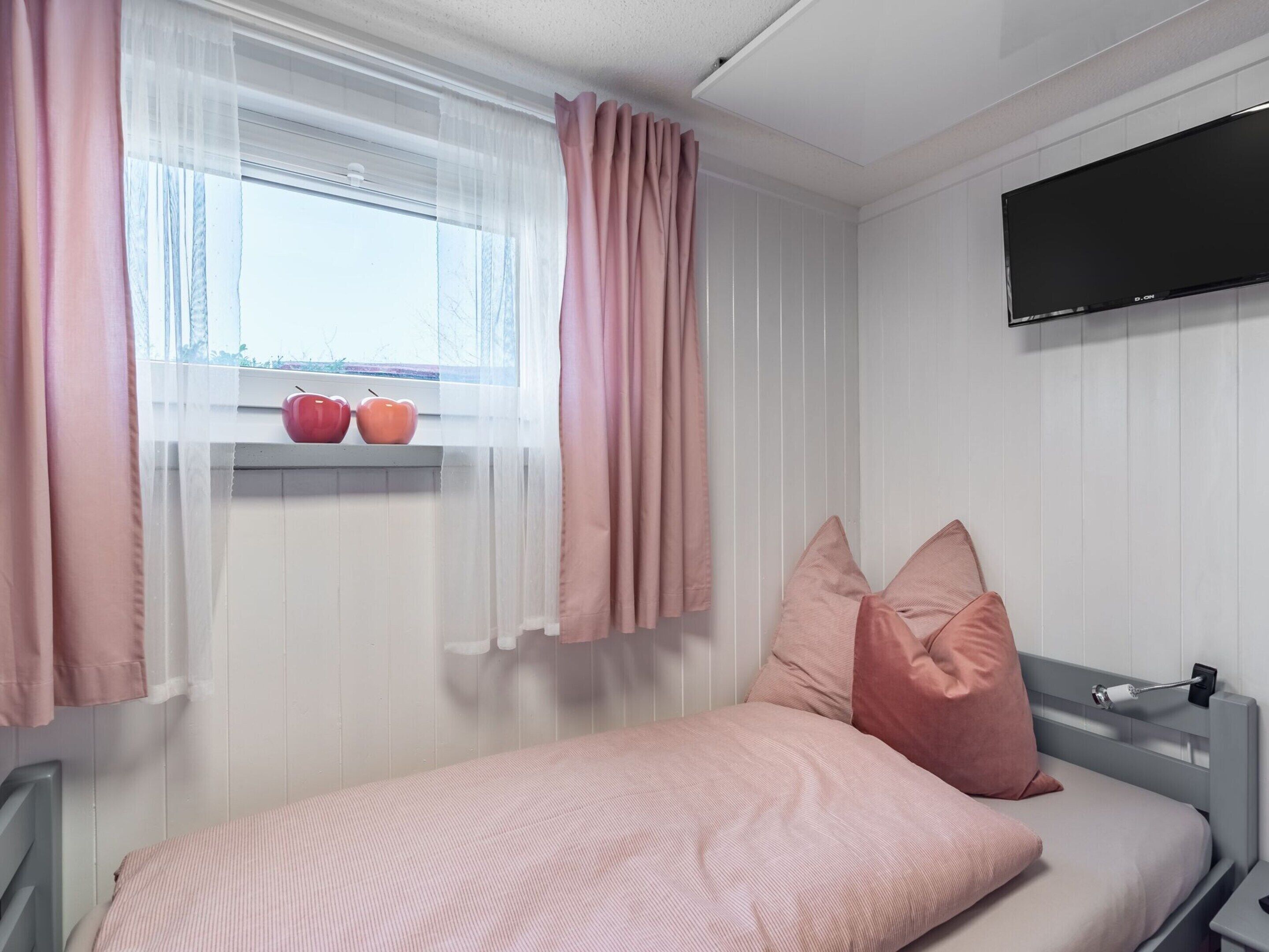 Un dormitorio con una cama, una ventana con cortinas translúcidas, un televisor montado y un estante con manzanas.