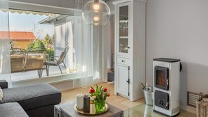 Een moderne woonkamer met een bank, een eettafel met stoelen, een glazen hanglamp, een witte kast en een houtkachel.
