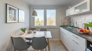 Een moderne keuken met een eettafel, stoelen, een hanglamp, een raam, keukenkastjes en een aanrecht met fruit.