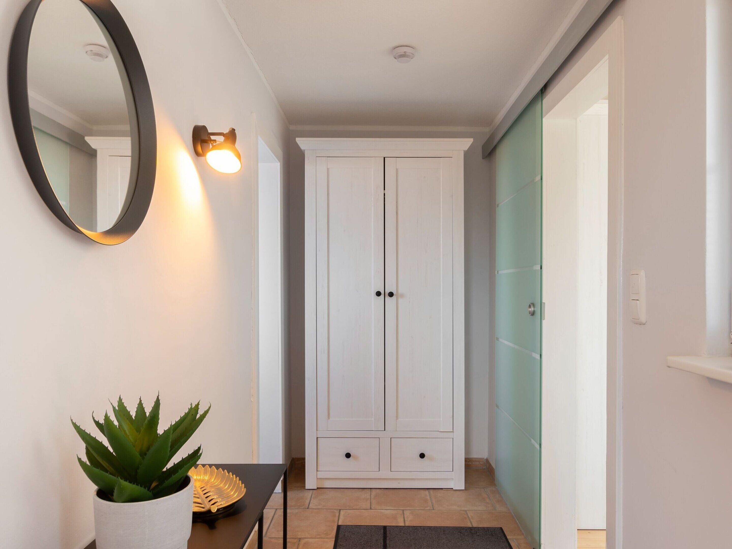Un couloir avec une armoire blanche, une plante en pot, un miroir et un luminaire fixé au mur.