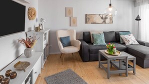 Een moderne woonkamer met een hoekbank, een witte fauteuil, een bijzettafeltje en een televisie aan de muur.