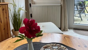 A room with a white sofa, a wooden table with a vase of red flowers, a potted plant, and a view of the outdoors through a window with curtains. - Holiday Home With Direct Access to the Beach (Ostseebad Baabe)