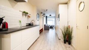 Una cocina moderna con armarios blancos, encimera negra y hervidor rojo. Hay un comedor con sofá y mesa, y un pasillo con una planta en maceta.